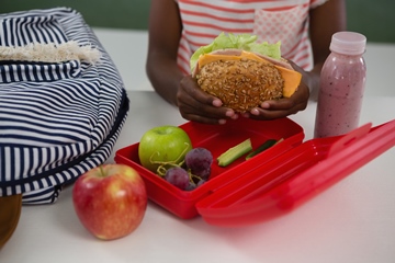 Schoolgirl holding a healthy sandwich in her hand with an open lunch box in front of her and a bottle of a strawberry smoothie