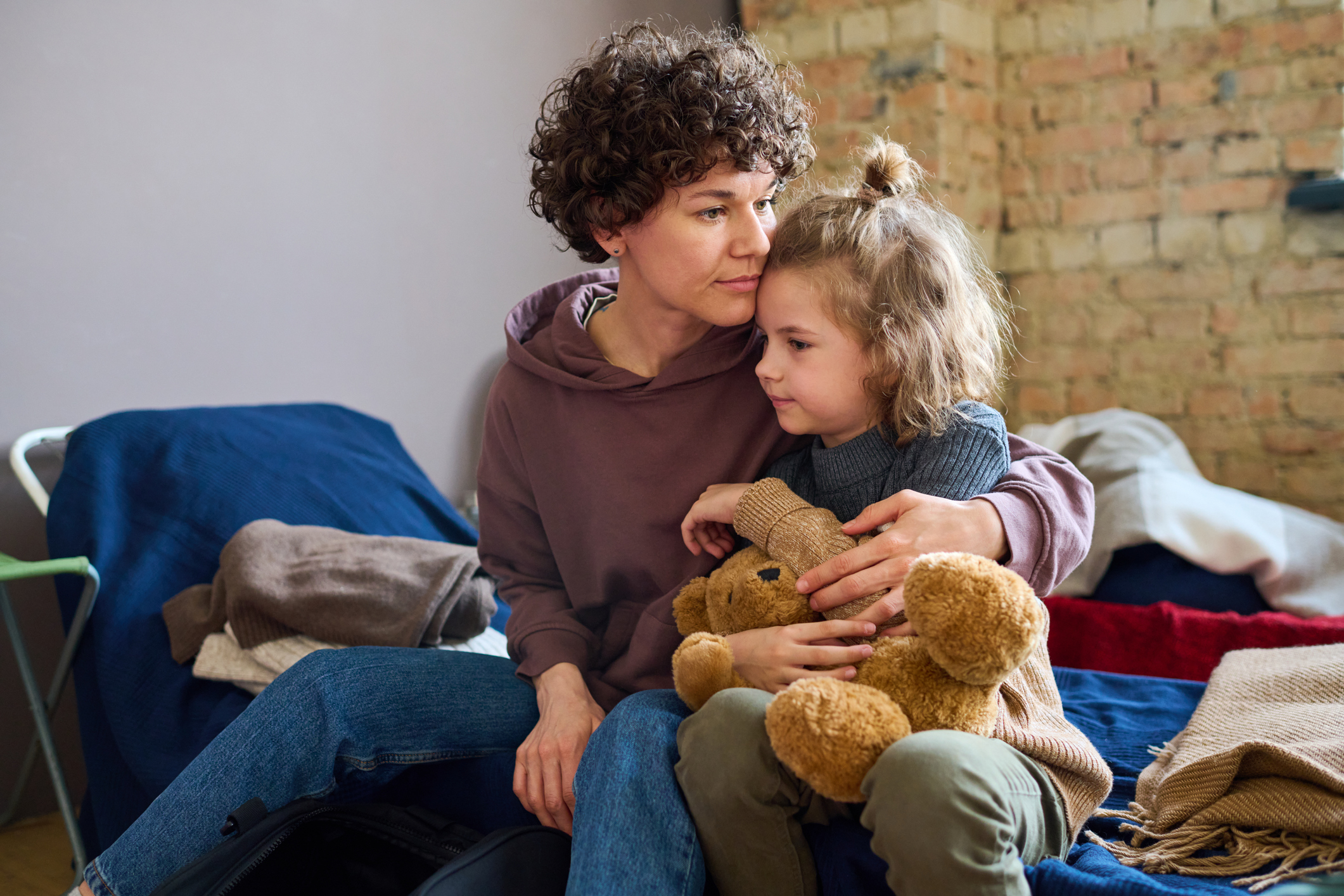 Young woman giving hug to her cute little son with brown soft teddybear while both sitting on sleeping place prepared for women in need